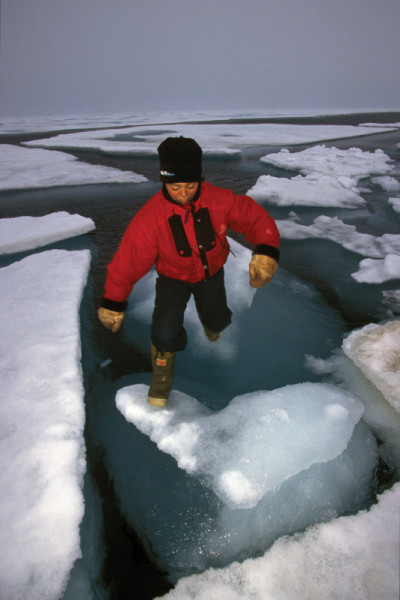 A boy in a red winter coat and black toque leaping across chunks of melting arctic sea ice, Nunavut, Canada.