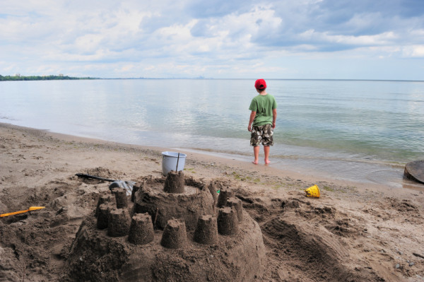 A young boy standing on the shores of Lake Ontario looks toward the skyline of Toronto after building a sandcastle. Mississauga, Ontario, Canada.
