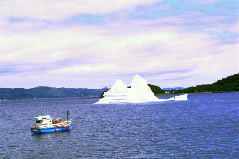 Iceberg from the Atlantic Ocean floats close to a fishing boat, off the coast of Newfoundland, Canada, with harbour shores in the background.
