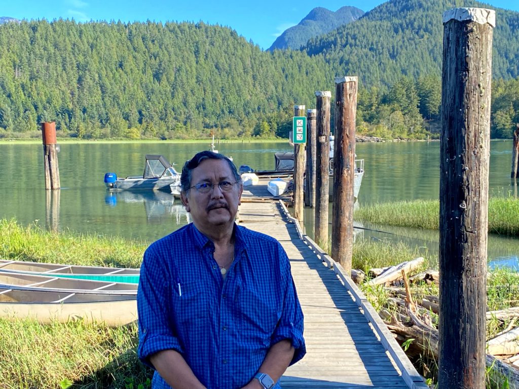 Vyras iš Katzie First Nation stovi saulėje prie ežero su kalnais tolumoje