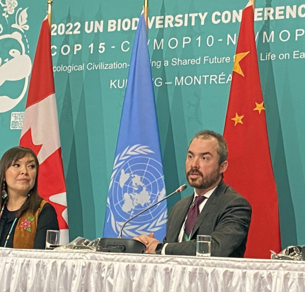 A woman and a man at a press conference table in front of flags from Canada, China and the United Nation