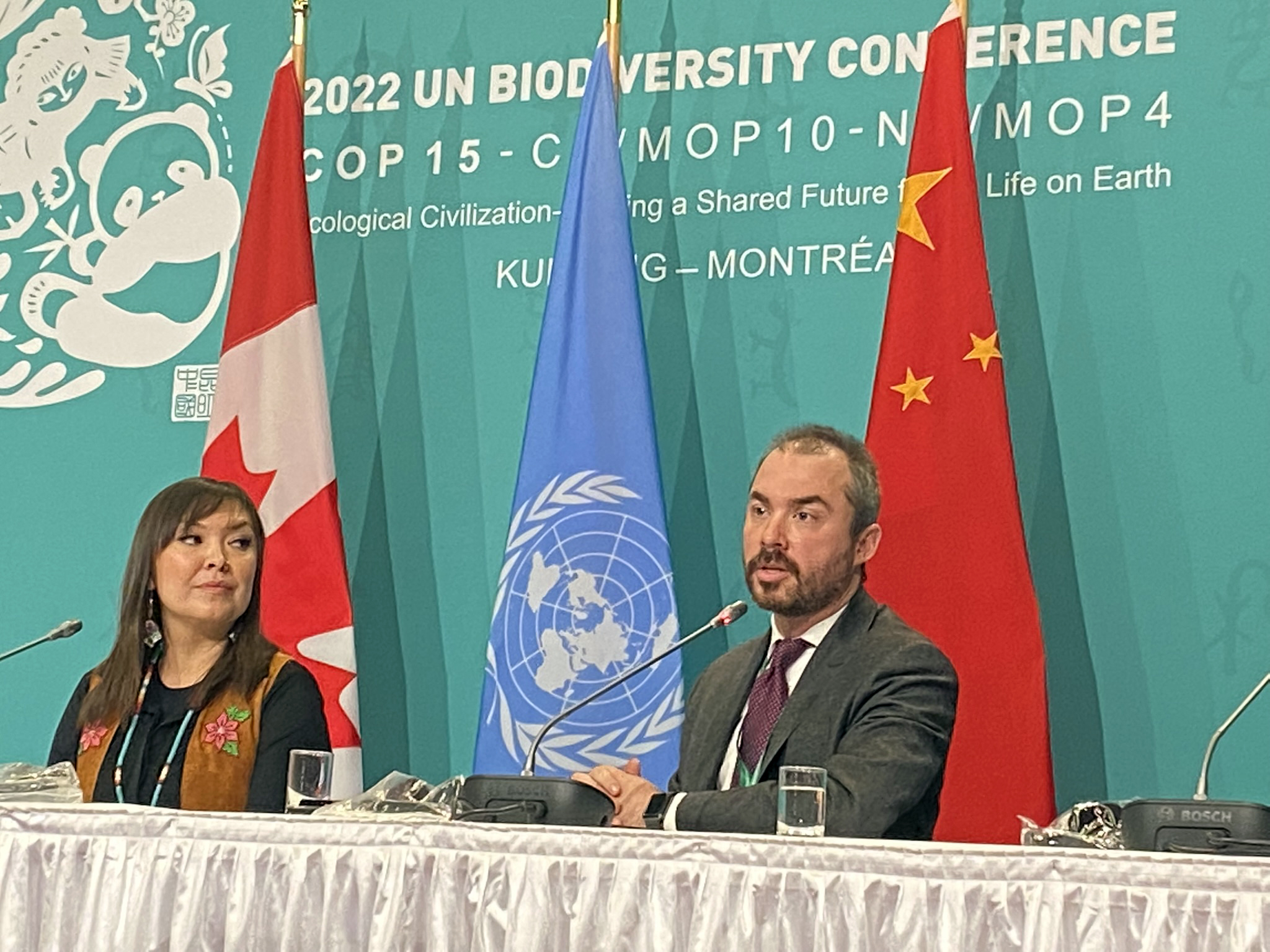 A woman and a man at a press conference table in front of flags from Canada, China and the United Nation