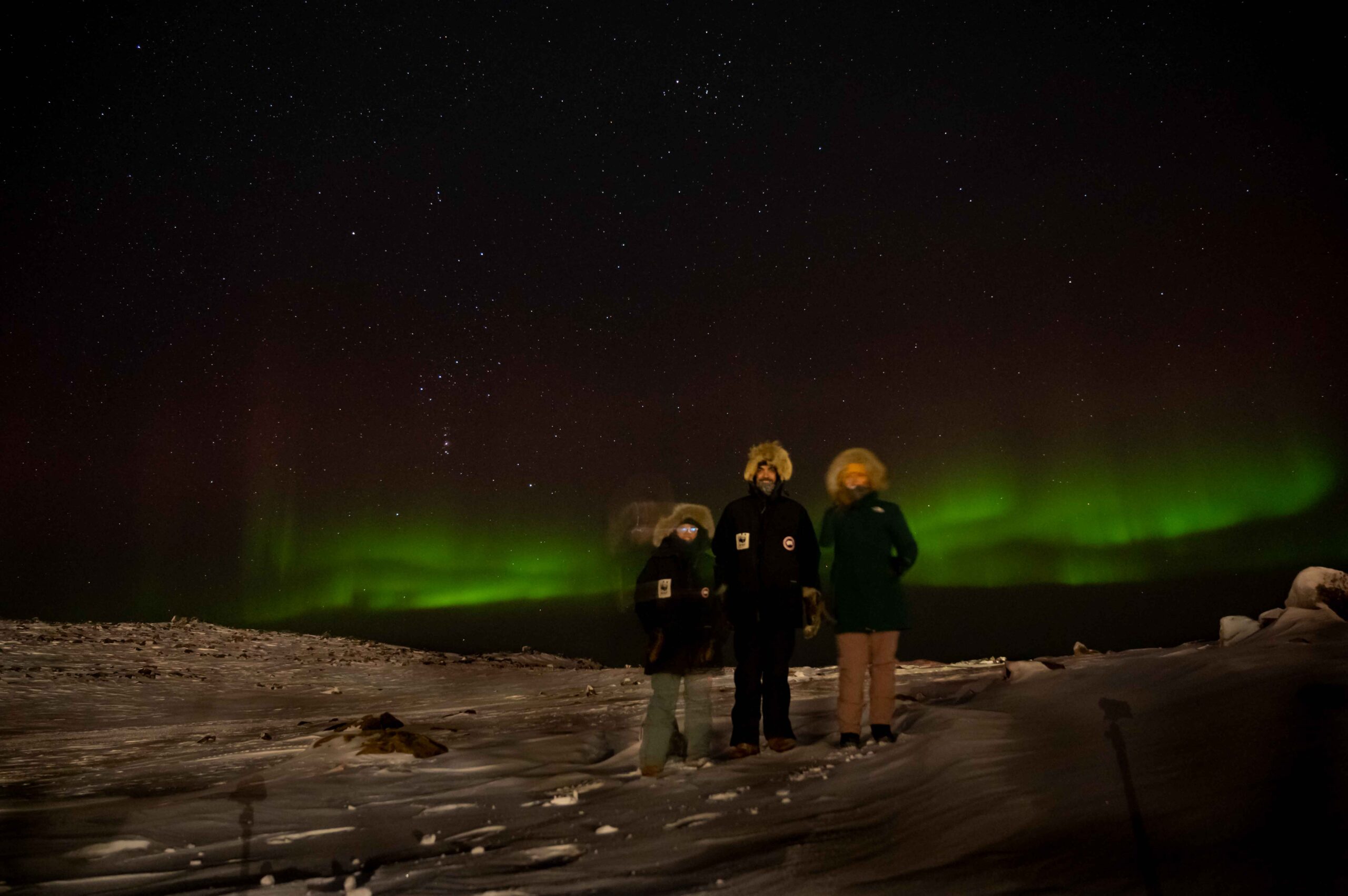 Three people in Arctic gear on the snow in front of a green Aurora Borealis