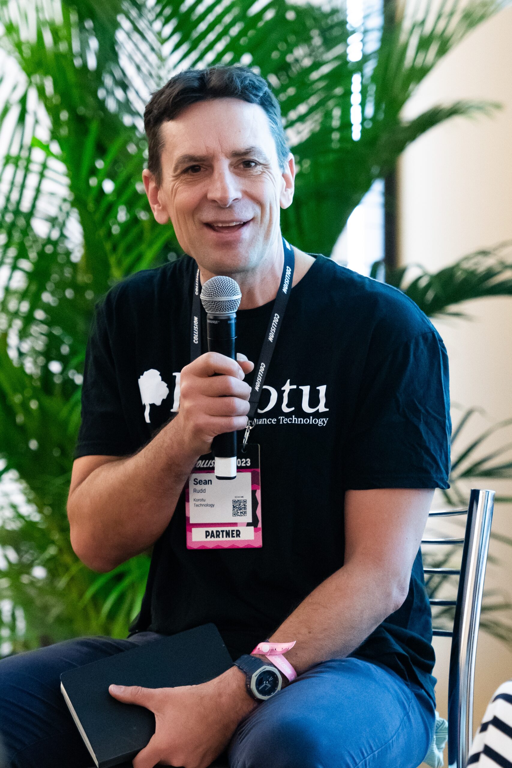 Man in black t-shirt, seated, holding a microphone 