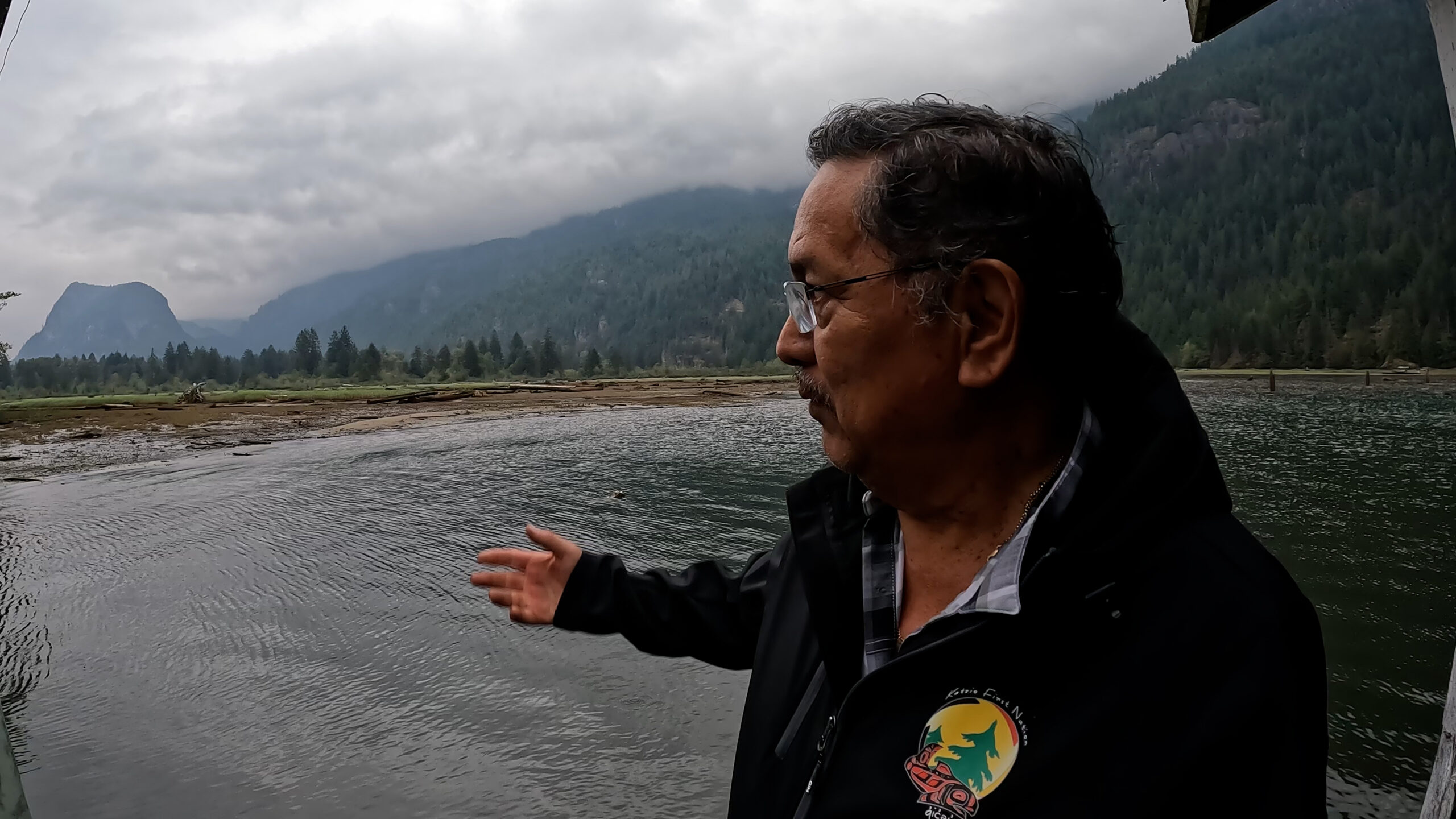 Katzie First Nation man pointing at a body of water with mountains in background