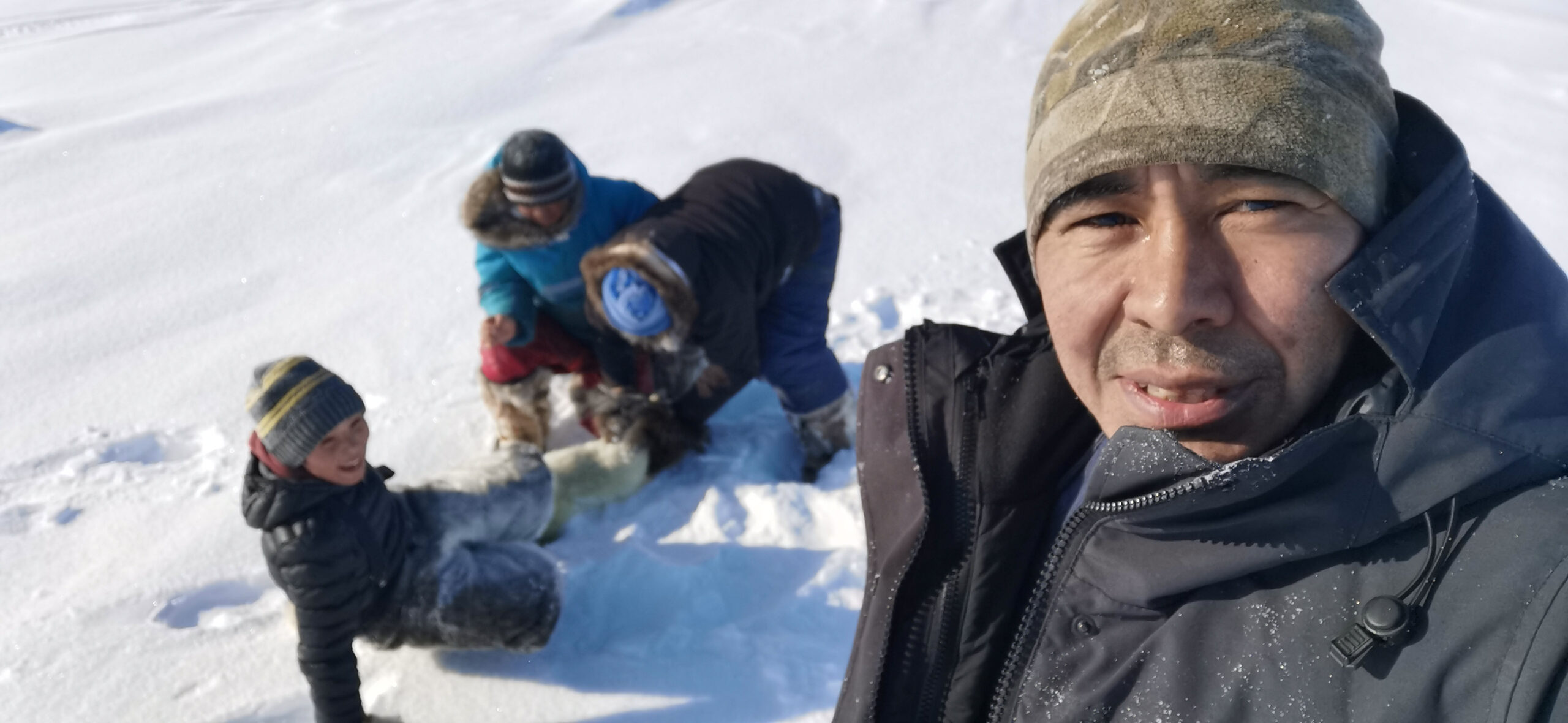 An Inuit man standing foreground with children behind him playing in the snow