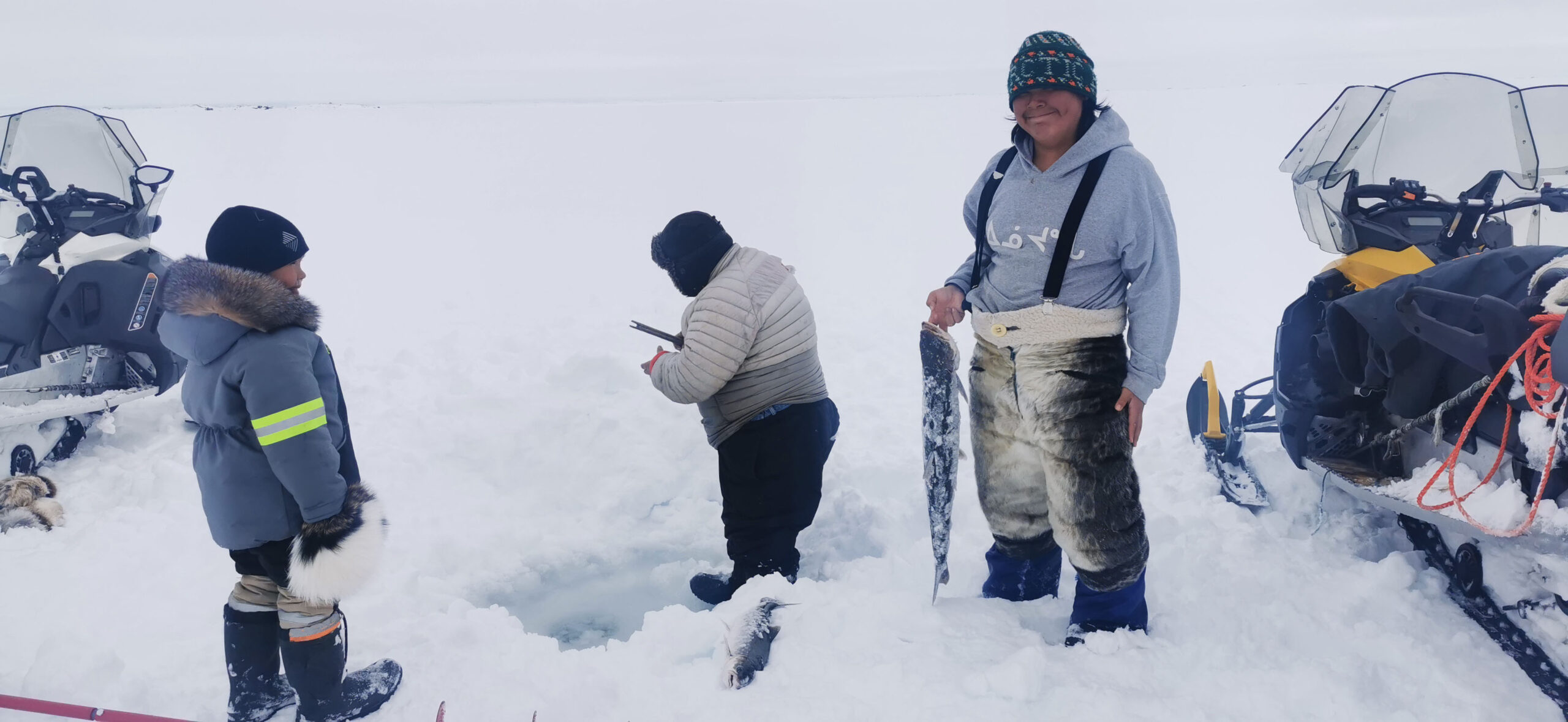 Scène de pêche sur glace inuite, autour d’un trou dans la banquise, trois personnes se tiennent debout et une jeune femme tient un poisson. 