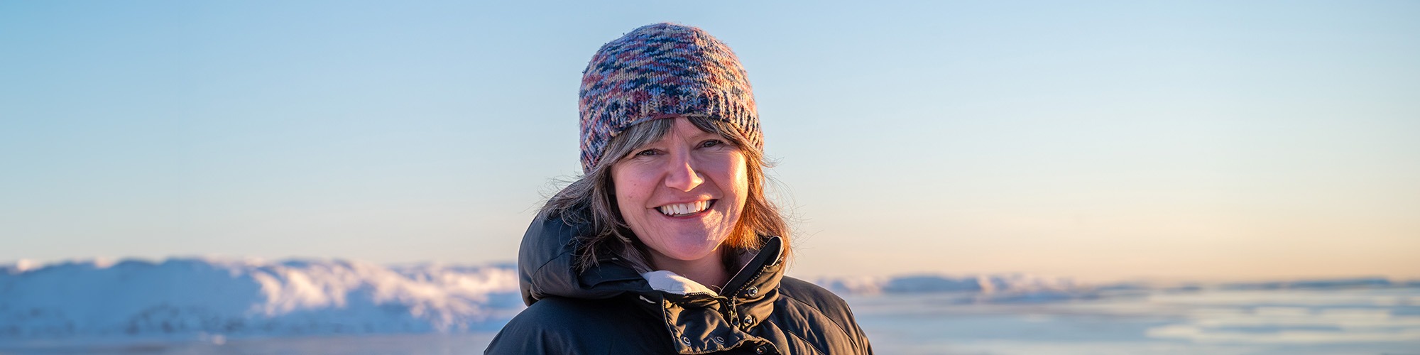 Woman in a toque smiling in the sunshine in front of an Arctic background