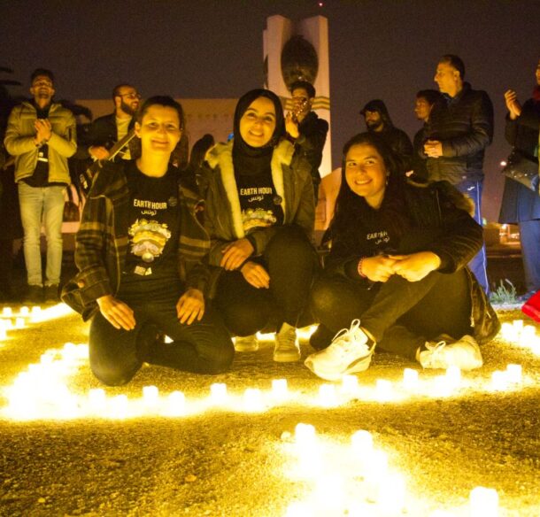 Three smiling young adults sit inside a circle of lights arranged on the ground outside at an evening gathering.
