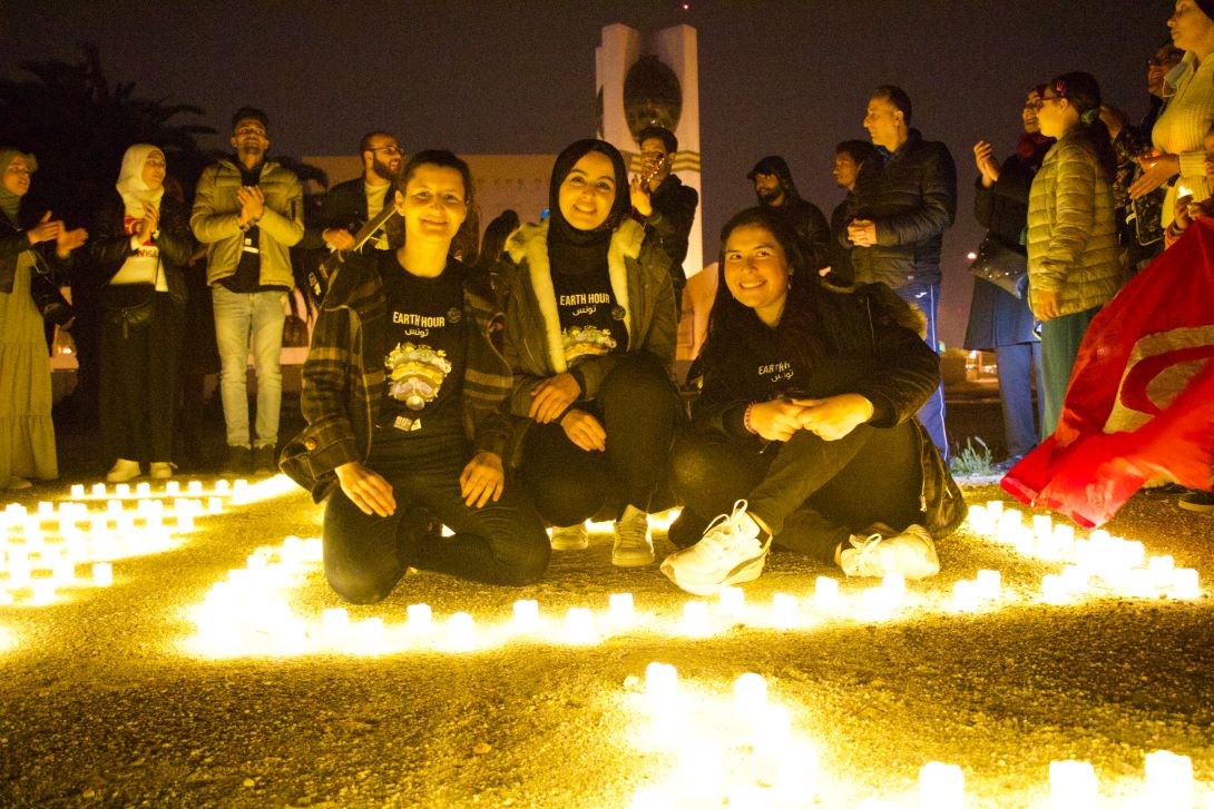 Three smiling young adults sit inside a circle of lights arranged on the ground outside at an evening gathering.