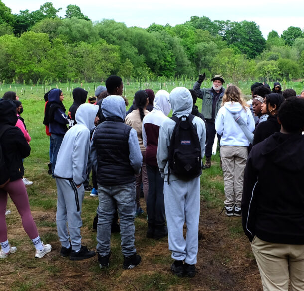 A man speaks to a group of teenage students in a meadow.