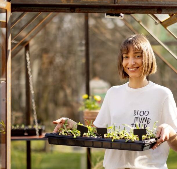 A smiling young adult carries a tray of seedlings from a small greenhouse into a yard on a sunny morning.
