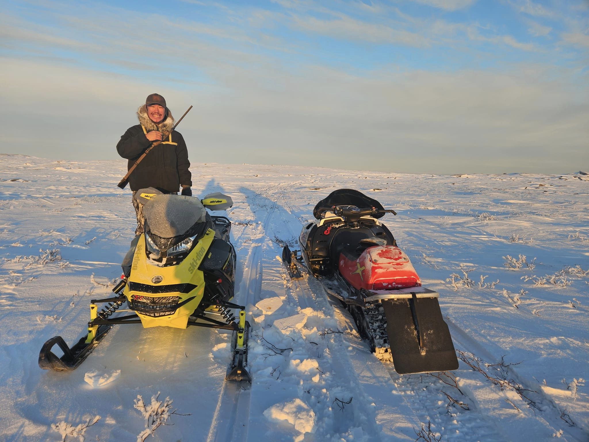 An Inuk man on a yellow snowmobile with a riderless red snowmobile beside him