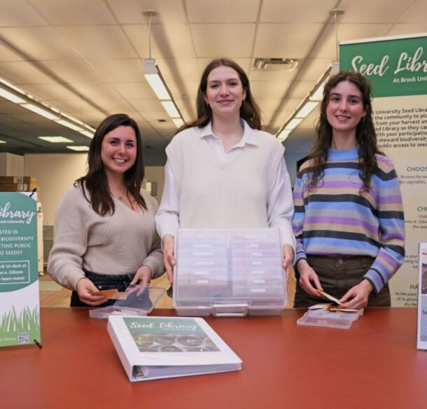 Three people holding containers of seed envelopes smile standing at a counter in a library beside three signs promoting the seed library.