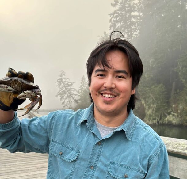 First Nations man in a denim shirt holding a crab and standing on a wooden bridge over water with a foggy forest behind him