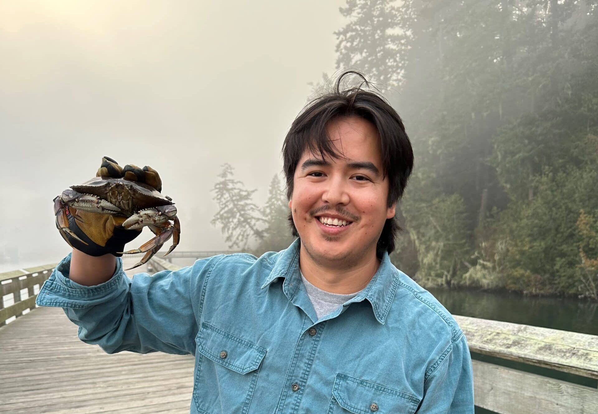 First Nations man in a denim shirt holding a crab and standing on a wooden bridge over water with a foggy forest behind him