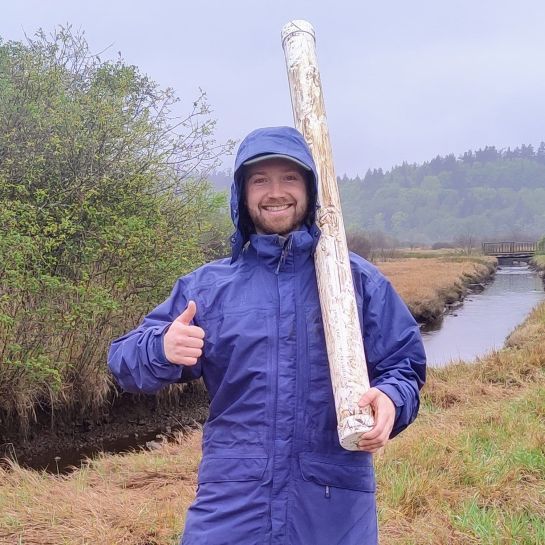 Cathal smiling and giving a thumbs up while holding a soil core in a long plastic sleeve, wearing a raincoat and standing beside a stream.
