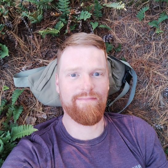 Close-up of Ryan lying on soil strewn with pine needs with his head resting on a small bag.