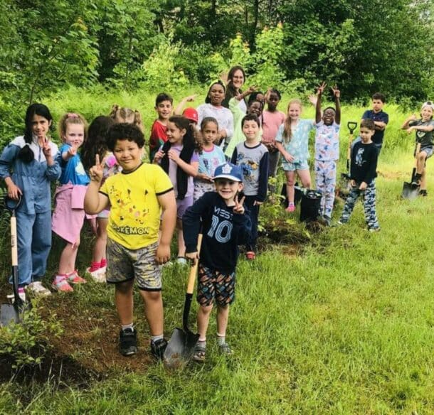 A group of 20 kids posing with shovels and small shrubs planted at the edge of a grassy field.