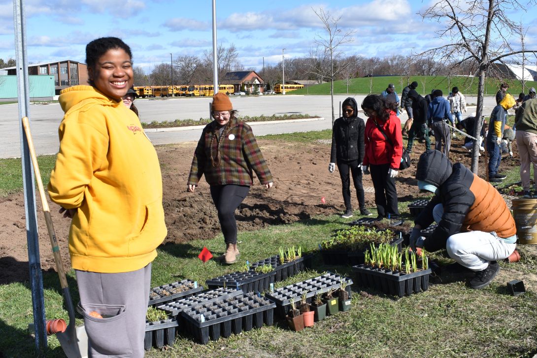A young adult smiling and standing beside a shovel in front of a dozen other young adults kneeling and standing around potted plants and bare soil outside on a sunny day.