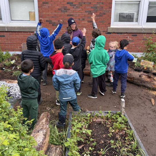 Eleven primary school children and one adult stand surrounded by gardens planted in raised beds and the ground. The children are facing the adult and three of them are raising their hands.