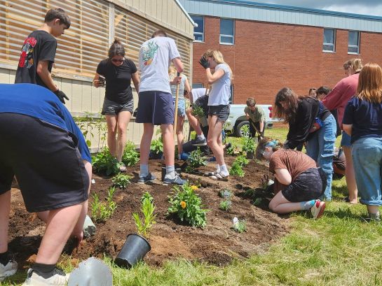 Young teens and adults dig holes and plant flowers in a garden bed beside a greenhouse and brick building.