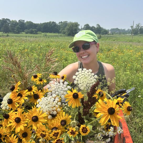 Isabel smiles while wearing a baseball cap and sunglasses as she stands in a field behind a bouquet of wildflowers.