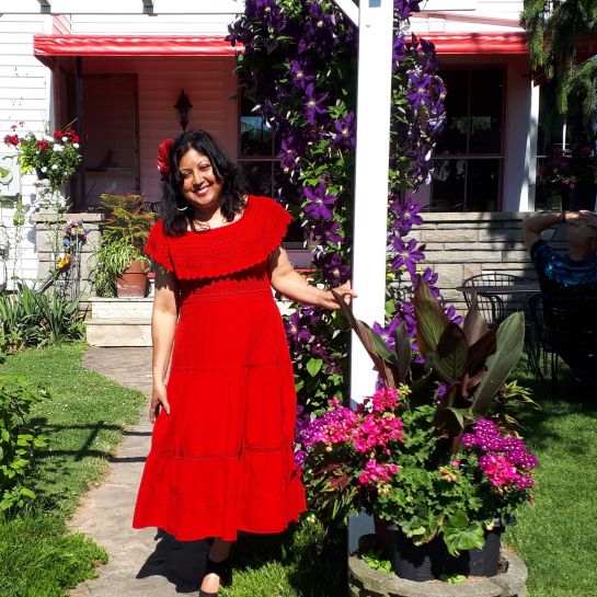 Juliet smiles at the camera wearing a red dress while standing beside flowering vines and potted plants in front of a house.