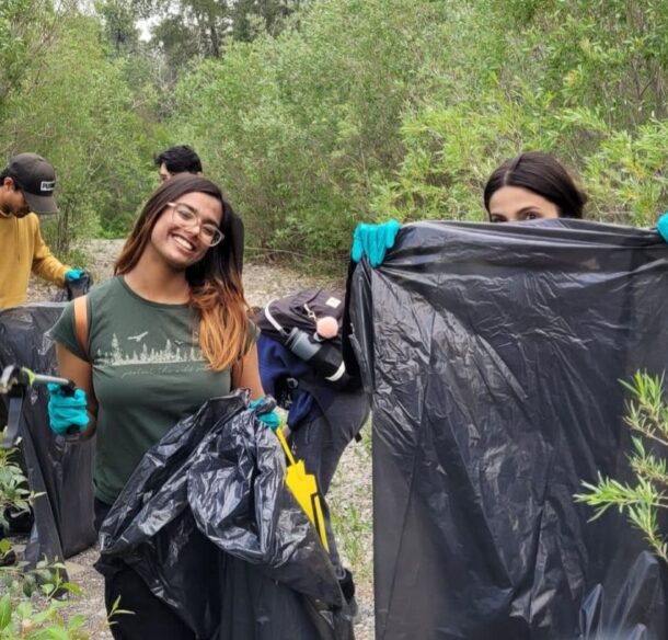 Four students with garbage bags and gloves standing among shrubs on a gravel beach. One student on the left, Srinidhi, smiles to the camera.