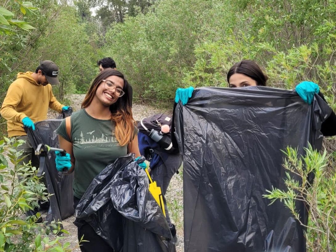 Four students with garbage bags and gloves standing among shrubs on a gravel beach. One student on the left, Srinidhi, smiles to the camera.